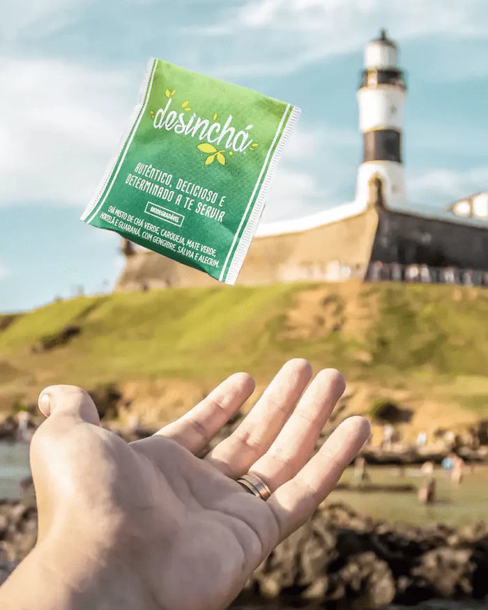 Sachê verde Desinchá lançado no ar sobre a mão, com o Farol da Barra, em Salvador, ao fundo