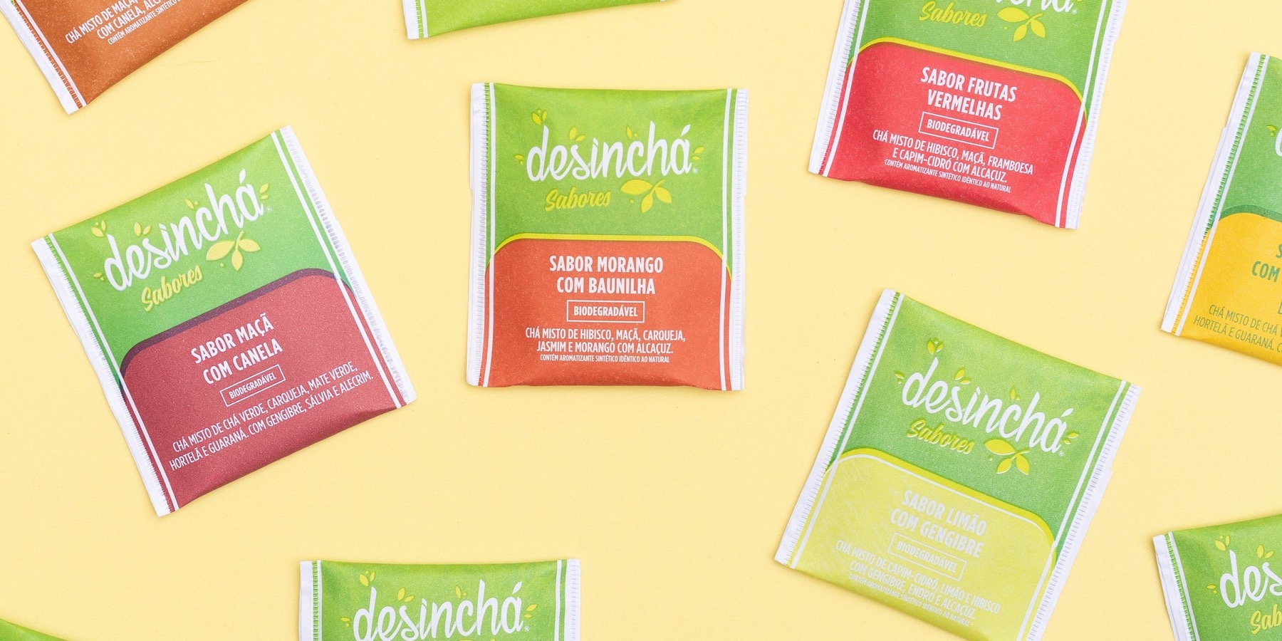 Sachês da linha Desinchá Sabores espalhados sobre fundo amarelo: maçã com canela, morango com baunilha, frutas vermelhas e limão com gengibre