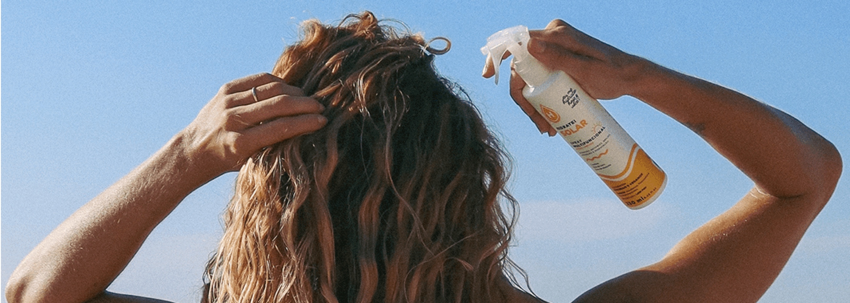 Mulher de costas, de cabelo ondulado, aplicando o Spray Multifuncional da Linha Solar da Hidratei contra o céu azul